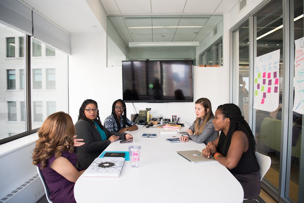 Five diverse women collaborating in a modern office meeting room, discussing projects.