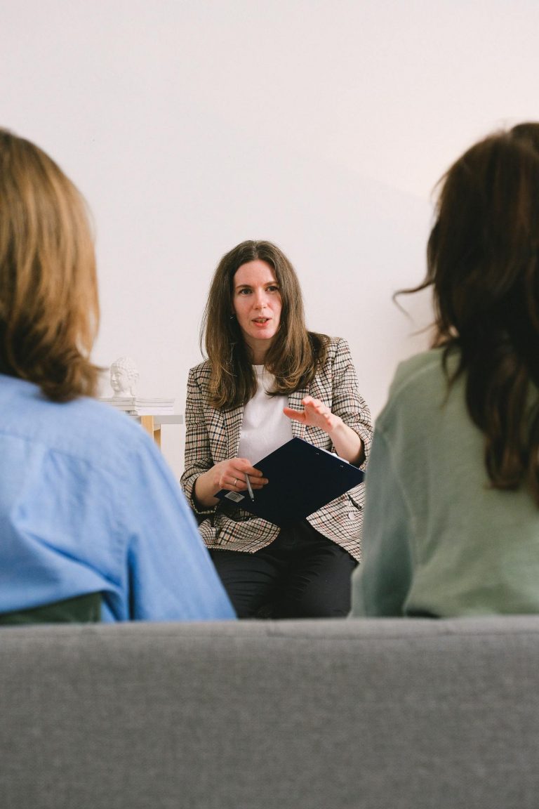 Pensive woman entrepreneur with brown hair in casual clothes sitting with clipboard and talking with unrecognizable clients in light room